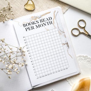 Könnte beinhalten: Ein offenes Notizbuch mit einer "Gelesene Bücher pro Monat"-Tabelle, goldenen und marmorierten Akzenten und einem Raster zur Verfolgung. Eine goldene Schere, ein herzförmiger Anhänger und das Wort "BUJO" sind ebenfalls sichtbar. Das Notizbuch ist von Stiften und dekorativen Elementen umgeben.