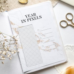 Könnte beinhalten: Aufgeschlagenes Notizbuch mit einer marmorierten Seite mit der Aufschrift "YEAR IN PIXELS". Die Seite enthält ein Raster zur Gewohnheitsverfolgung mit Kontrollkästchen und Linien für Notizen. Eine goldene Schere und eine goldene herzförmige Verzierung sind ebenfalls zu sehen.