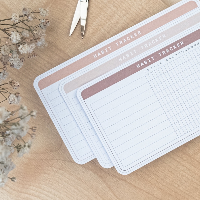 May include: Three habit tracker cards with a pink, light pink, and brown border. The cards have a grid pattern and the text "HABIT TRACKER" at the top.