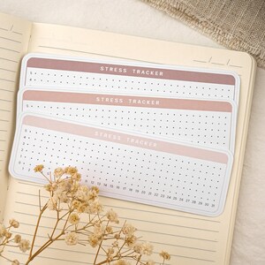 May include: Three pink and brown stress tracker stickers with a grid of dots and the text "STRESS TRACKER" printed on each sticker. The stickers are on a lined notebook page.