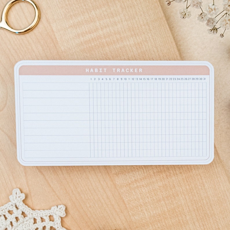 May include: A white habit tracker with a pink header and the text "HABIT TRACKER" printed on it. The tracker has 31 columns for each day of the month and rows for different habits.