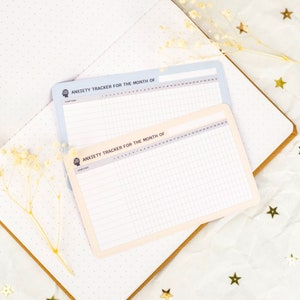May include: Two printable anxiety trackers for the month of. The trackers are on white paper with a light blue and cream background. The trackers have a grid pattern and a space for writing the start date.