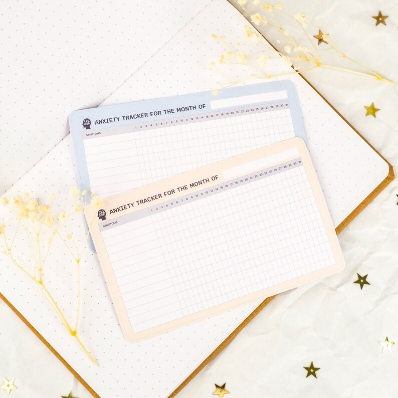 May include: Two printable anxiety trackers for the month of. The trackers are on white paper with a light blue and cream background. The trackers have a grid pattern and a space for writing the start date.