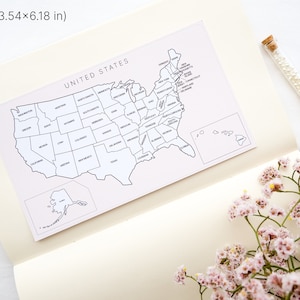 May include: A white and black map of the United States, measuring 3.54 x 6.18 inches, is displayed on a cream-colored notebook. The map is labeled "UNITED STATES" and includes state outlines. A glass vial of pearls and pink flowers are also present.
