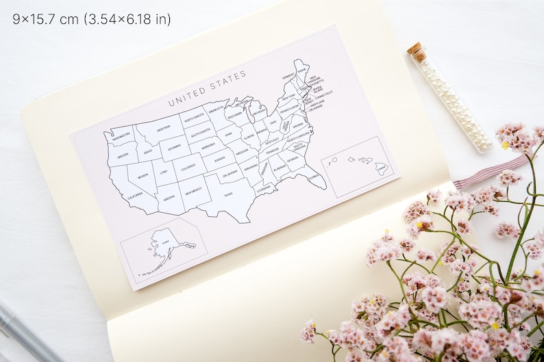 May include: A white and black map of the United States, measuring 3.54 x 6.18 inches, is displayed on a cream-colored notebook. The map is labeled "UNITED STATES" and includes state outlines. A glass vial of pearls and pink flowers are also present.