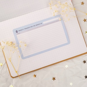 May include: A blue and white anxiety tracker for the month of with a grid for tracking symptoms. The tracker is on a page with a dotted pattern.
