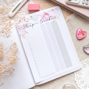 Op de afbeelding: Open notitieboek met een bloemenontwerp en de woorden "Sleep tracker". De pagina's bevatten een slaapschema met ruimte voor datum, tijd en slaapscore. Andere briefpapierartikelen, waaronder een rekenmachine en gum, zijn zichtbaar.