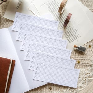 May include: A collection of white monthly habit tracker inserts with grid lines, laid out on an open brown notebook. Two rolls of decorative washi tape and loose pages of text are in the background. A small glass bottle with glitter is also present.
