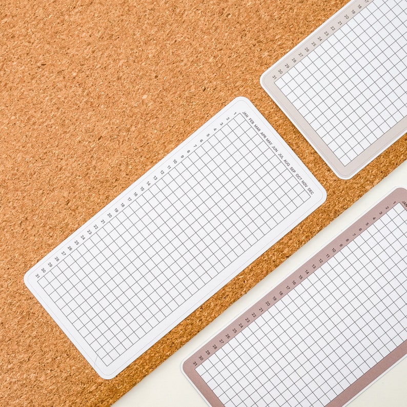 May include: Two white gridded sheets of paper with month names printed on the top sheet. The sheets are on a brown corkboard background.