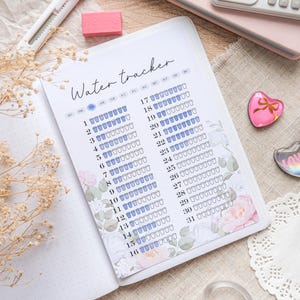 Può includere: Un diario aperto con una pagina "Water tracker", con un design floreale e un layout di calendario per monitorare l'assunzione di acqua. La pagina presenta icone di piccole tazze blu. Sono visibili altri articoli di cancelleria.