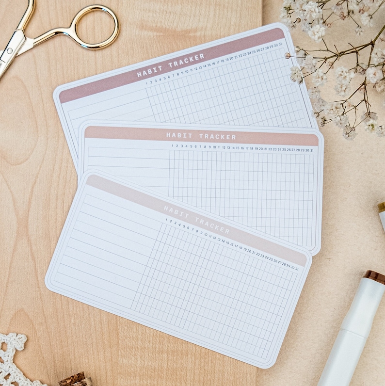 May include: Three white habit tracker cards with a light brown and pink gradient border. Each card has a grid with numbers 1 through 31 at the top and lines down the side. The text "HABIT TRACKER" is printed at the top of each card.