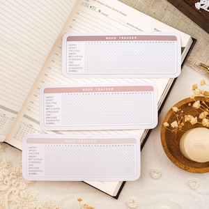 May include: Three pink and white mood trackers with a grid of 31 squares. Each tracker has a list of moods: happy, upset, excited, angry, motivated, stressed, sad, anxious, exhausted, and normal.