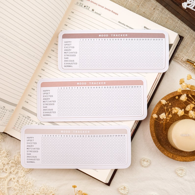 May include: Three pink and white mood trackers with a grid of 31 squares. Each tracker has a list of moods: happy, upset, excited, angry, motivated, stressed, sad, anxious, exhausted, and normal.