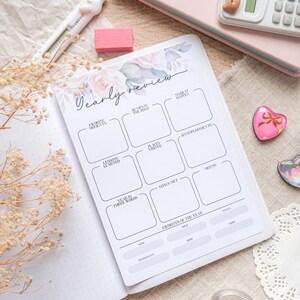 May include: An open planner with the words "Yearly Review" and floral accents. The planner has sections for "Favorite Moments," "Lessons Learned," and "Goals Met." A pink eraser, a white calculator, and other stationery items are nearby.