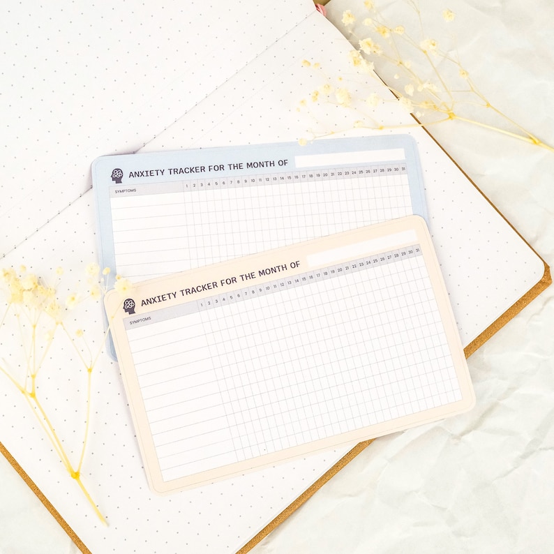 May include: Two printable anxiety trackers, one in light blue and one in cream, with the text "Anxiety Tracker for the Month of" and "Symptoms" at the top. The trackers have a grid layout with numbered days of the month and spaces for recording symptoms.