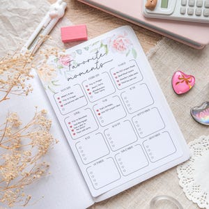 May include: An open planner with the words "Favorite moments" and floral accents. The planner has monthly sections with checkboxes. Other items include a pink notebook, calculator, eraser, and decorative heart-shaped magnets.