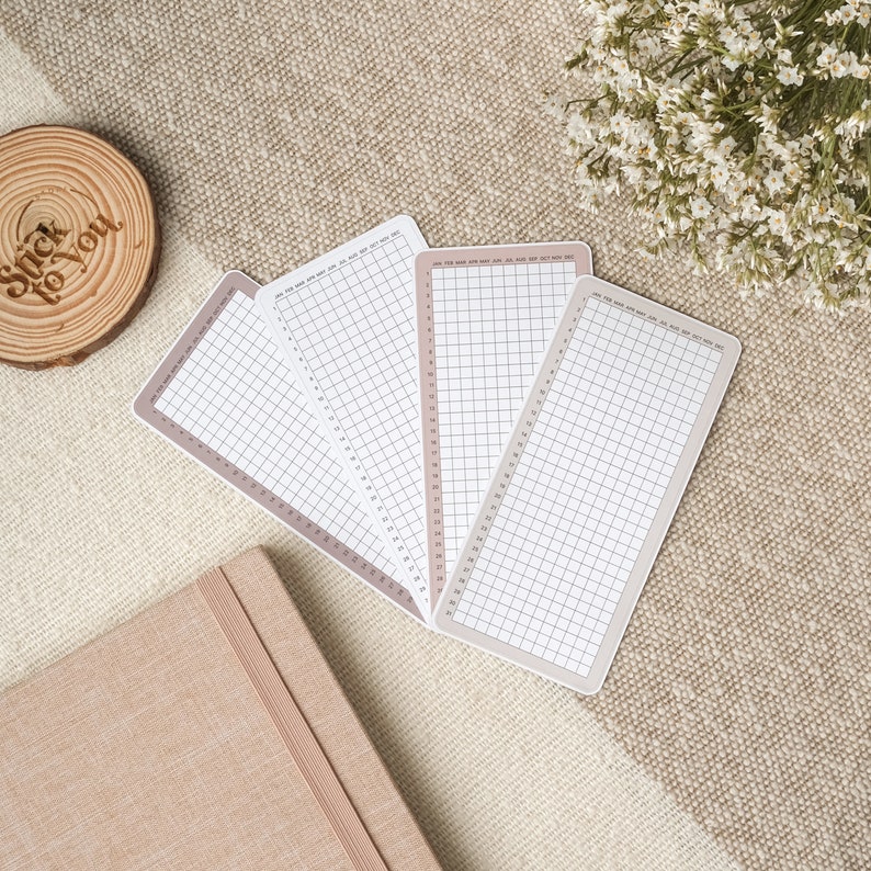 May include: Four white gridded notepads with a light brown border. The notepads are arranged in a fan shape on a beige textured surface.