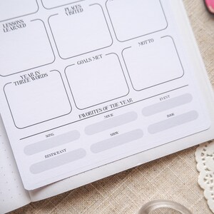 May include: A white planner page with sections for reflection, including "Lessons Learned," "Places Visited," and "Favorites of the Year." The page features a grid layout with black text and lines, designed for journaling and goal setting.