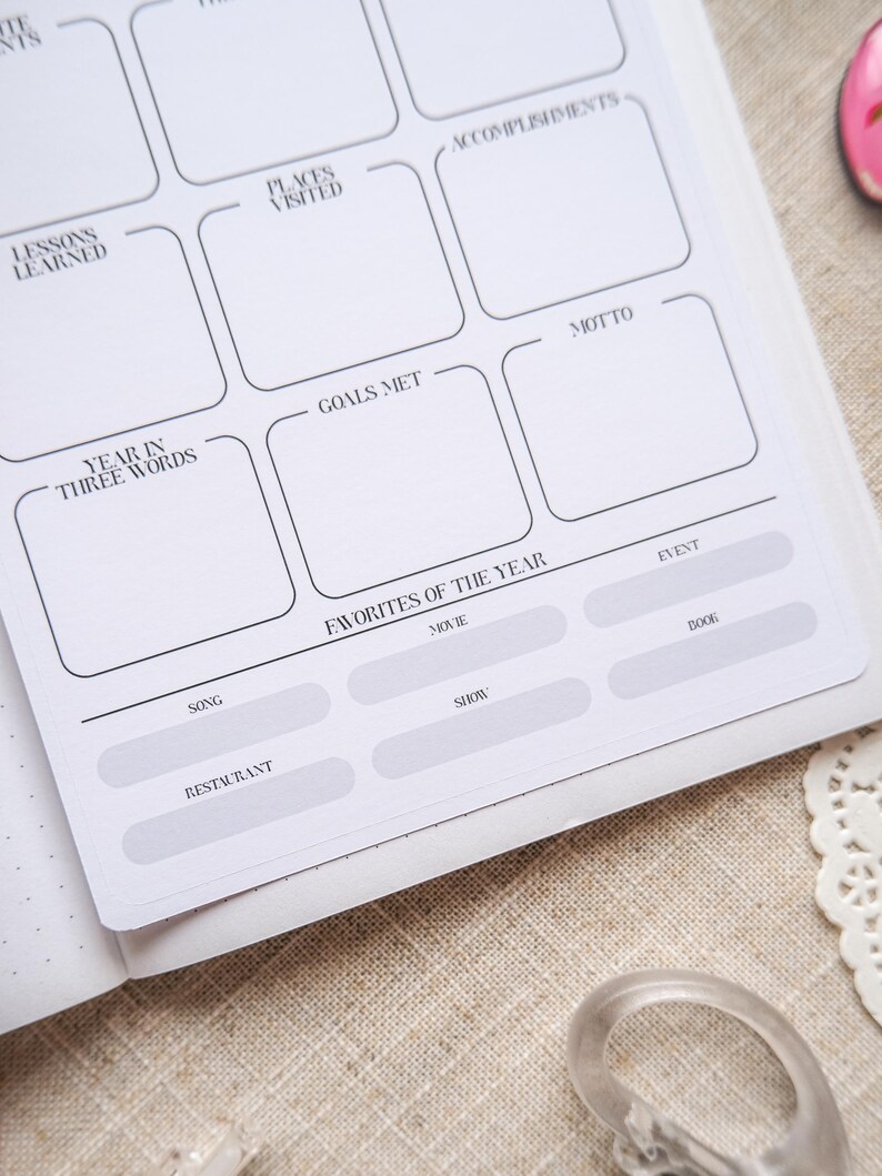 May include: A white planner page with sections for reflection, including "Lessons Learned," "Places Visited," and "Favorites of the Year." The page features a grid layout with black text and lines, designed for journaling and goal setting.
