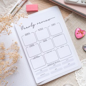 May include: A white planner with the title "Yearly Review" in cursive script. The planner includes sections for "Favorite Moments," "Bumps in the Road," and "Accomplishments." A pink eraser and a bunny-shaped cutter are visible.