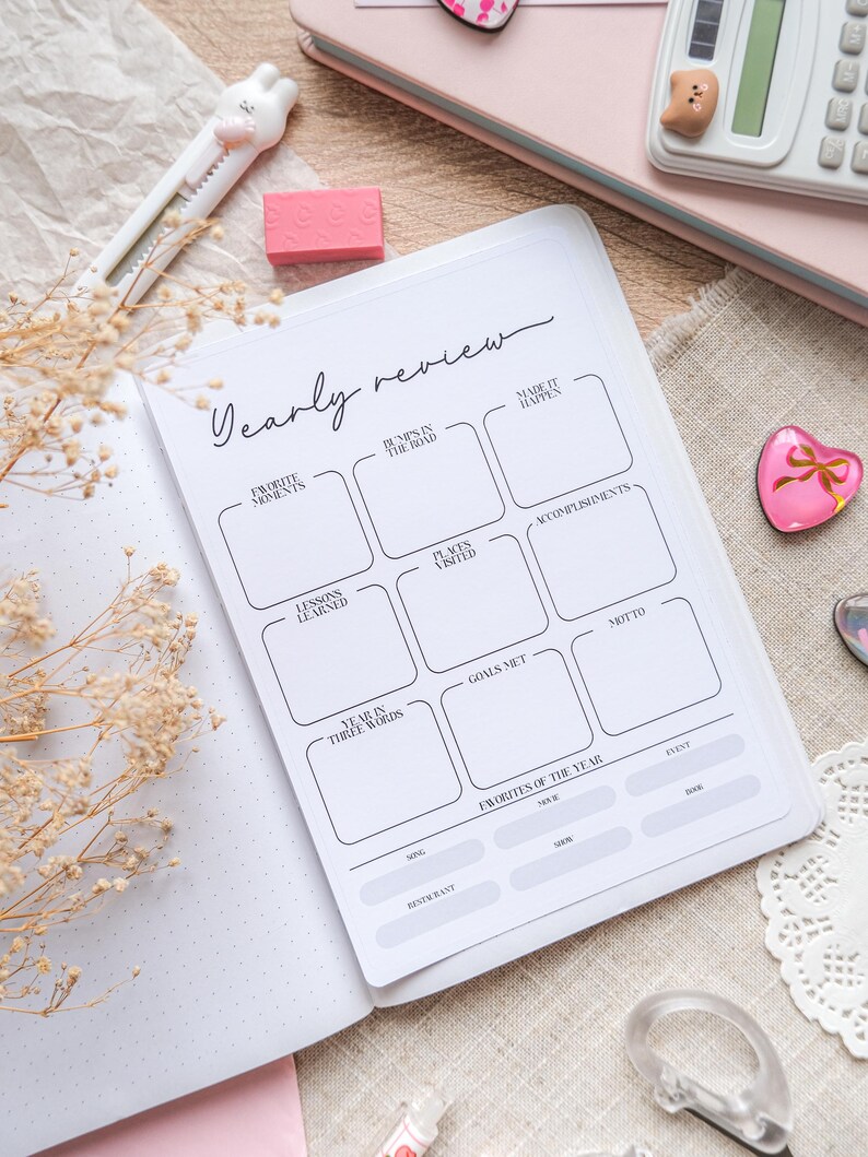 May include: A white planner with the title "Yearly Review" in cursive script. The planner includes sections for "Favorite Moments," "Bumps in the Road," and "Accomplishments." A pink eraser and a bunny-shaped cutter are visible.