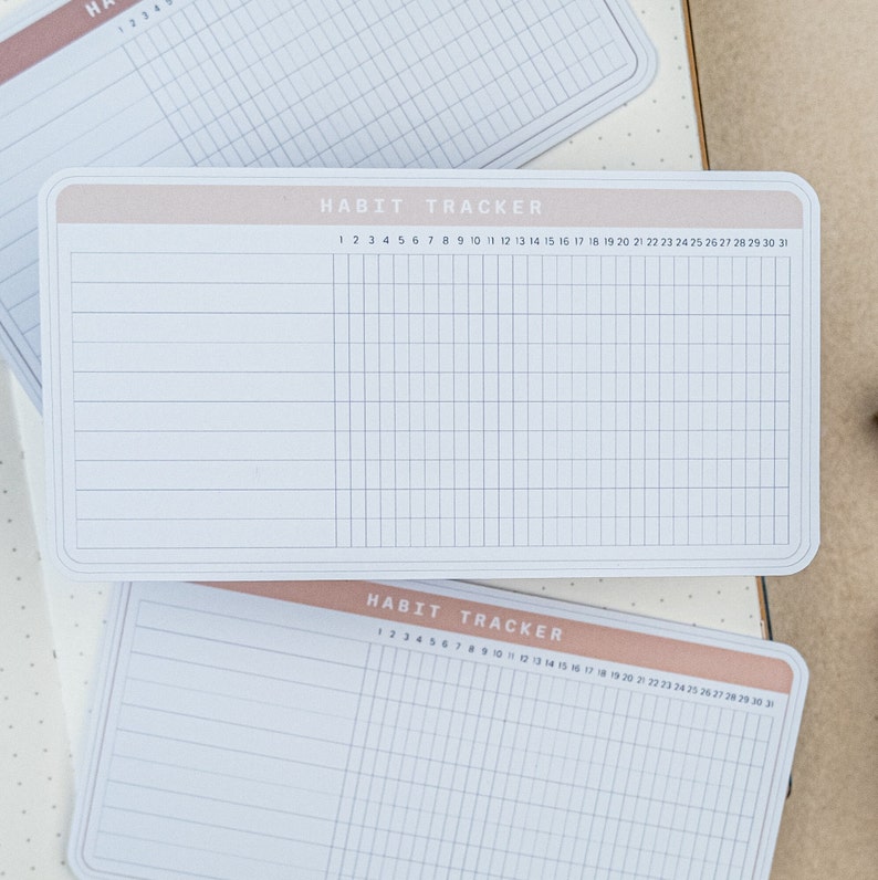 May include: Two white habit tracker sheets with a light pink header. The header says "Habit Tracker" and has the numbers 1 through 31 printed on it. The sheets are divided into 31 columns and 10 rows.