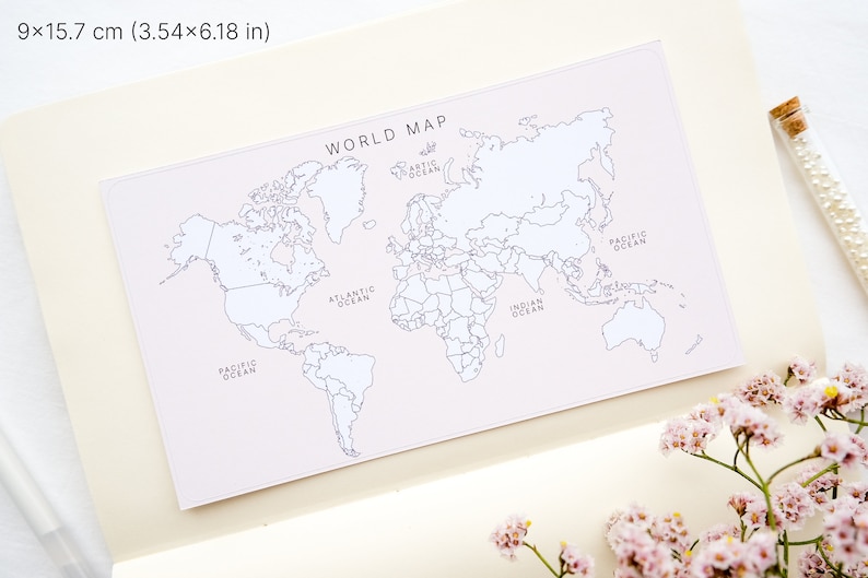 May include: A rectangular world map print, 3.54 x 6.18 inches, with continents and oceans labeled in black on a pale pink background. The words "WORLD MAP" are at the top. The map is set on a cream-colored notebook page.