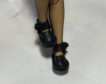 May include: Black doll shoes with a Mary Jane style, featuring a strap with a button closure. The shoes are made of a textured material and are worn with fishnet stockings. The shoes are small and appear to be for a doll.