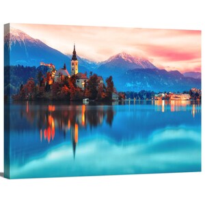 May include: A picturesque view of a church on a small island in a lake, with snow-capped mountains in the background and a colorful sunset sky reflected in the water.
