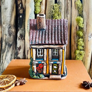 Könnte beinhalten: Ein kleines, handbemaltes Keramikhaus mit gelber Fassade, braunem Dach und grünen Verzierungen. Das Haus hat zwei Fenster, eine Tür und einen Schornstein. Es steht auf einem braunen Buch mit einer dekorativen Orangenscheibe und Sternanis.