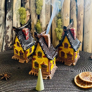 Może przedstawiać: Trzy żółte ceramiczne domki z czarnymi dachami i czerwonymi ceglanymi kominami. Domki są palnikami kadzideł z palącym się stożkiem kadzidła w środkowym domku. Domki stoją na brązowej plecionej macie z suszoną plastrą pomarańczy i anyżem gwiazdkowym.