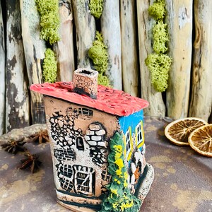 Puede incluir: Una figurita de casa de cer&aacute;mica pintada a mano con un techo rojo, paredes azules y una chimenea. La casa tiene un exterior detallado con mamposter&iacute;a, ventanas y un peque&ntilde;o &aacute;rbol.
