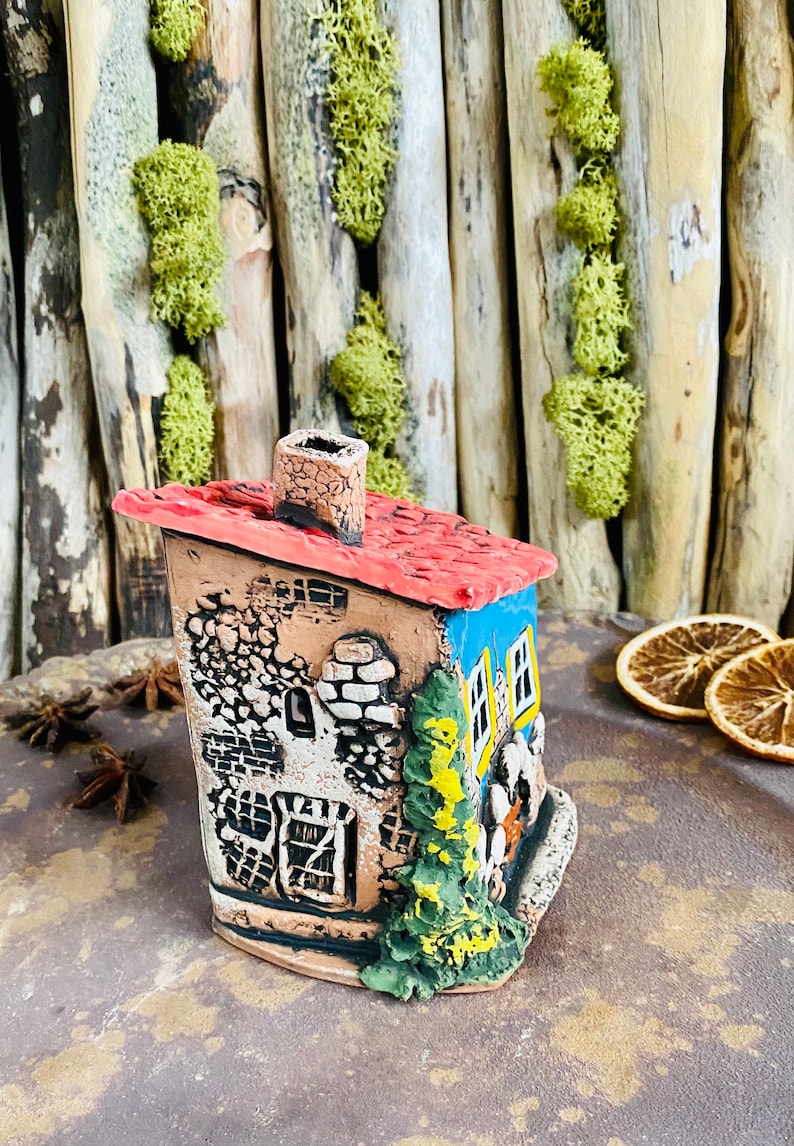 Puede incluir: Una figurita de casa de cer&aacute;mica pintada a mano con un techo rojo, paredes azules y una chimenea. La casa tiene un exterior detallado con mamposter&iacute;a, ventanas y un peque&ntilde;o &aacute;rbol.