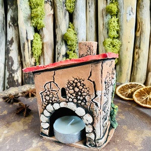 Puede incluir: Un peque&ntilde;o portavelas de cer&aacute;mica con forma de casa con un techo rojo y una vela de t&eacute; blanca en su interior. La casa tiene un exterior texturizado y una peque&ntilde;a ventana.