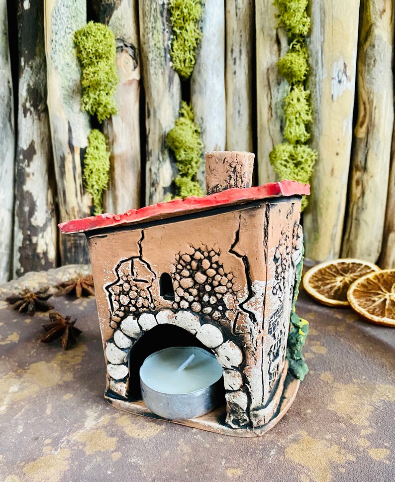 Puede incluir: Un peque&ntilde;o portavelas de cer&aacute;mica con forma de casa con un techo rojo y una vela de t&eacute; blanca en su interior. La casa tiene un exterior texturizado y una peque&ntilde;a ventana.