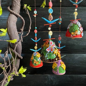 Può includere: Campanelli eolici in ceramica colorata con corpi a forma di campana, adornati con motivi floreali e a farfalla. I campanelli presentano accenti rossi, verdi, viola e gialli, sospesi a fili di perline. Un ramo decorativo è visibile.
