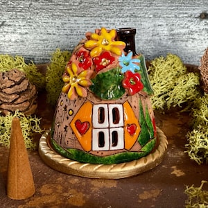 Peut inclure: Une maison de fée en céramique avec une cheminée, ornée de fleurs colorées et d'une fenêtre en forme de cœur. La maison repose sur une base dorée, entourée de mousse, d'une pomme de pin et de cônes d'encens.
