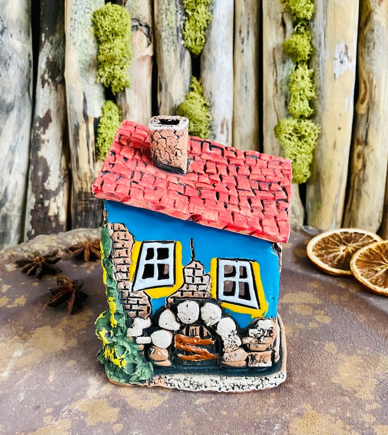 Puede incluir: Una peque&ntilde;a casa de cer&aacute;mica azul y roja con chimenea, ventanas y fachada de piedra. La casa est&aacute; decorada con pintura amarilla y tiene una base verde con hojas.