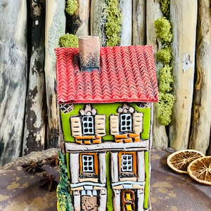 Op de afbeelding: Een handgeschilderd keramiek huisbeeldje met een groen lichaam, een rood dak en witte versieringen. Het huis heeft twee ramen op elke verdieping en een kleine deur. Het beeldje staat op een bruin oppervlak.