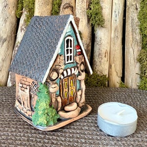 May include: A ceramic house shaped candle holder with a teal green door and a gray roof. The house has a small tree and a brick wall on the side. A white tea light candle is included.