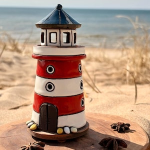 May include: A ceramic lighthouse with a red and white striped body, a black roof, and a small door. The lighthouse sits on a wooden base with decorative stones. The background shows a sandy beach and the ocean.