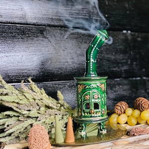 Peut inclure: Brûle-encens en céramique verte en forme de petite maison avec une cheminée. Le brûleur a un design détaillé avec des accents dorés et est posé sur une surface en bois avec des cônes d'encens et des fleurs séchées.