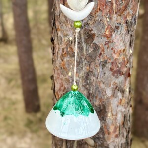 Op de afbeelding: Een handgemaakte windgong met een natuurlijke uitstraling. Het heeft een witte, klokvormige bodem met een groene, gespikkelde bovenkant. Bovenop hangen een witte halve maan en een kleine, ronde steen met groene kralen en natuurlijk touw.
