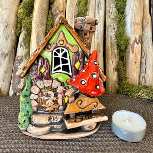 Può includere: Una casetta di fata in ceramica con tetto verde, muri in pietra marrone e un fungo rosso e bianco. La casa ha una piccola porta, una finestra e un camino. Una candela da tè si trova davanti alla casa.