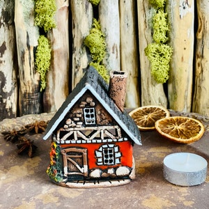 Pode incluir: Uma pequena casa de cerâmica vermelha e marrom com uma chaminé e um telhado branco. A casa tem uma porta e janelas, e é decorada com detalhes brancos e marrons. A casa está sentada em uma superfície marrom com fatias de laranja secas e uma vela de chá.