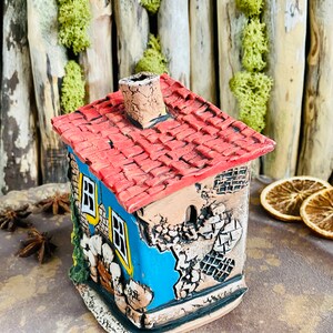 Puede incluir: Una peque&ntilde;a casa de cer&aacute;mica azul con un techo de tejas rojas y una chimenea. La casa tiene una ventana y una puerta, y est&aacute; decorada con detalles blancos y amarillos. La casa est&aacute; sentada sobre una superficie marr&oacute;n.