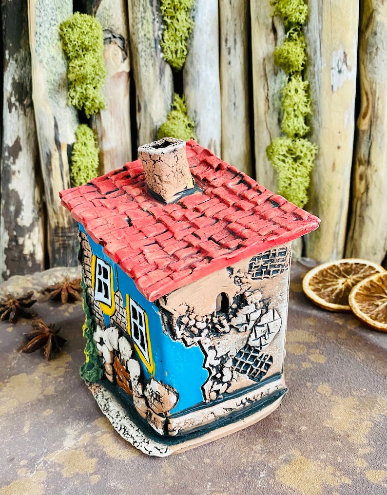 Puede incluir: Una peque&ntilde;a casa de cer&aacute;mica azul con un techo de tejas rojas y una chimenea. La casa tiene una ventana y una puerta, y est&aacute; decorada con detalles blancos y amarillos. La casa est&aacute; sentada sobre una superficie marr&oacute;n.