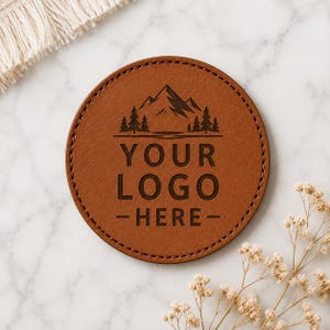 May include: A round brown leather coaster featuring a dark engraved mountain and pine tree graphic above the text YOUR LOGO HERE. The coaster has visible stitching along the circular edge and a smooth, matte finish on the surface.