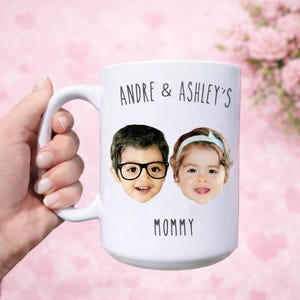 May include: White ceramic mug featuring the faces of two children, one wearing glasses and the other a headband. The mug is inscribed with "Andre & Ashley's" above and "Mommy" below the images. The mug is held in a hand.