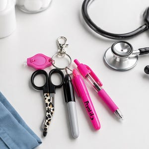 Op de afbeelding: Een verzameling medische hulpmiddelen en accessoires, waaronder een stethoscoop, roze pennen, een zwarte marker, een klein roze lampje en een schaar met een luipaardprint. De items zijn gerangschikt op een wit oppervlak.